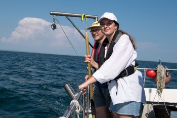 A female and male student stand at the edge of a boat, holding onto the railings, looking out to sea.