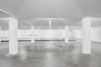 A open room with white walls and white pillars, featuring a fountain pouring from the ceiling onto the concrete floor.