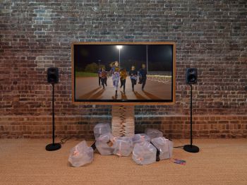 A large TV displaying 6 young adults on a running track, running towards the screen. It’s dark but the track is lit by floodlight.