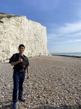 Student consultant Ryota is wearing a black t shirt and jeans, and giving the camera a thumbs-up. They are stood in front of a white cliff on a stony beach