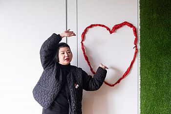 photograph of kar-man au in a black quilted jacket with three badges on the left lapel, she has her hands above her head gesturing towards a big read heart on a wall, made of tissue paper.