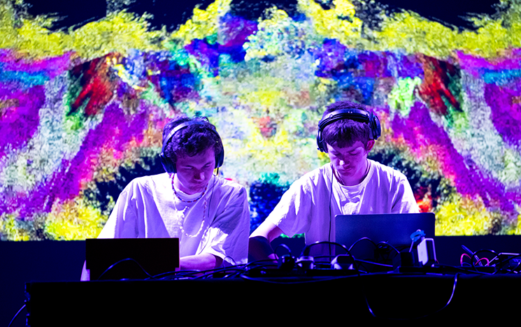 Two students performing a DJ set in front of a colourful background screen.