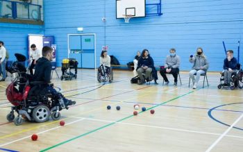 In the Sports Centre, a student on a chair is throwing a small blue ball at other blue and red balls on the floor in front of he. Next to her, four students on wheelchairs or chairs wait their turn. Opposite a man on wheelchair encourages her to play