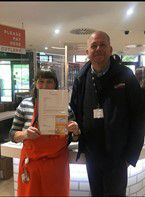 woman on a red apron holding a certificate next to taller bold man wearing a dark anorak