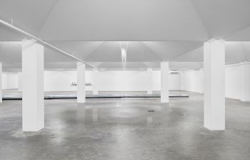 A open room with white walls and white pillars, featuring a fountain pouring from the ceiling onto the concrete floor.