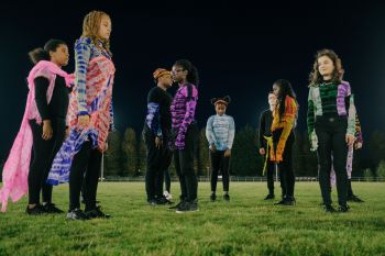 9 young adult females standing still on a flood lit grass pitch at night. All wearing black trousers with colourful tie dyed tops.