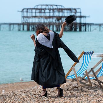 Finalist dressed with the gown and holding the mortarboard in the right hand on Brighton beach, wit the sea and the West Pier is on the background