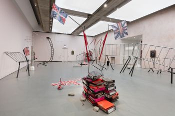 A room filled with contrasting objects; warped metal barriers and structures stand elevated, a pile of orange, pink and black books, a warped ladder and structures with White, Red and Blue fabric resembling the Union Jack flag.