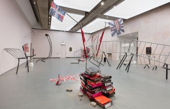 A room filled with contrasting objects; warped metal barriers and structures stand elevated, a pile of orange, pink and black books, a warped ladder and structures with White, Red and Blue fabric resembling the Union Jack flag.