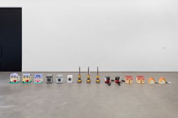 15 miniature toy objects all laid out in a row with the exact spacing between them. Order from left to right is; 3 larger houses, 3 washing machines, 3 yellow Dyson hovers, 2 red Henry Hoovers, 2 medium houses, 2 bungalows.