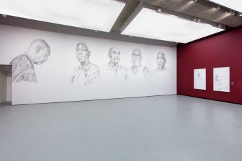 A white wall covered with 5 large-scale wall drawings of 2 women and 3 men affected by the Windrush scandal.  Faces convey a mix of strength and vulnerability, intricate charcoal strokes create a compelling and emotive portrayal of the subjects.