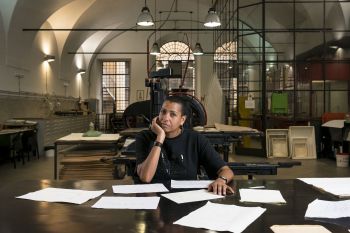 Helen Cammock sits as a desk surrounded by papers. She is in a large industrial studio with three large arched windows behind her.
