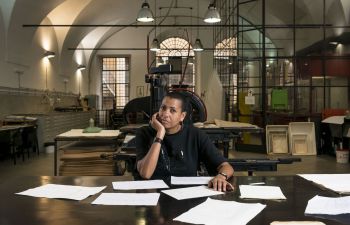 Helen Cammock sits as a desk surrounded by papers. She is in a large industrial studio with three large arched windows behind her.