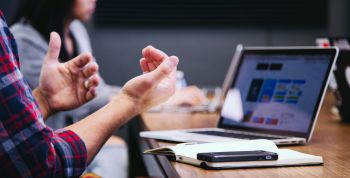 Person gesturing in front of a laptop, phone, and notebook