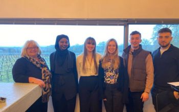 An employee of Ridgeview Wine Estate (the lady on the left) and five University of Sussex students stand in front of a large window with a view of the vineyard.