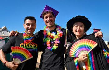 A photo of three people wearing graduation gowns with rainbow fans, and rainbow University of Sussex t-shirts. There is a blue sky in the background.