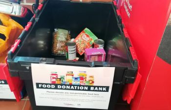 Inside a black donation crate, there are a few non-perishable food items, including canned goods and a packet of pasta. A sign in front of the crate reads, “FOOD DONATION BANK - Please donate any non-perishable food here.”