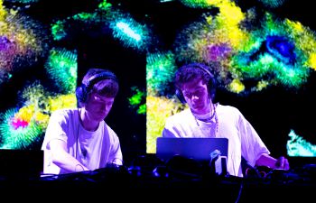 Music duo 'eyeveyes' performing on stage. Behind them is a multicolour pattern projected on a screen.