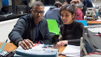 Two individuals engaged in an electronics project in a classroom, with other students working in the background