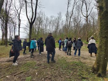 A group of individuals on a nature walk in a wooded area during the colder months, with leafless trees and a cloudy sky above