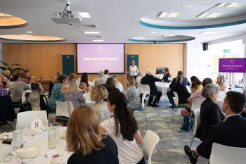 A conference room with people seated at round tables facing a presenter in front of a screen displaying ‘Welcome to Sussex.’