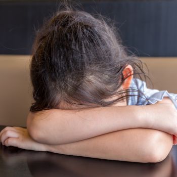 A child with dark hair resting their head in the arms on a table