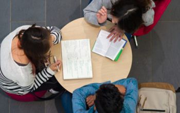 Overhead view of three individuals seated around a round table with open notebooks, engaged in study or discussion.