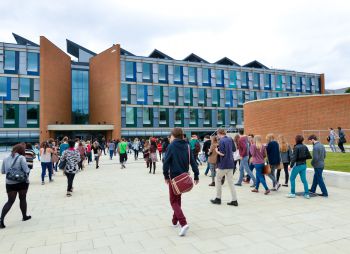 A modern educational building with a large glass entrance and brick facade. Numerous students are walking in front of the building, suggesting a busy campus environment.