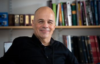 David Ruebain with a black shirt sitting in front of a bookcase