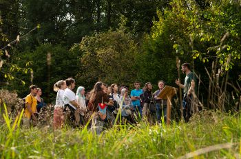 A group of students stand in a lush green area, in front of woodland, and listen to an academic