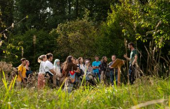 A group of students stand in a lush green area, in front of woodland, and listen to an academic