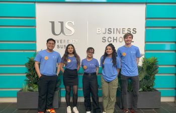 Five individuals stand in front of a wall with the Business School logo printed on it. The individuals are wearing blue t-shirts with a circular yellow badges on the chest.