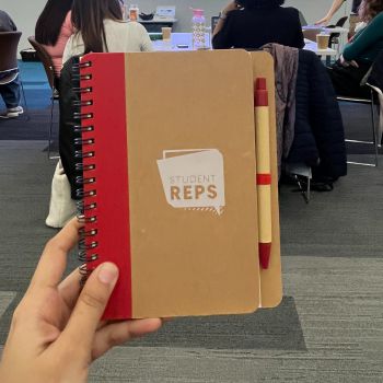 A hand holding up a notebook branded with the student rep scheme