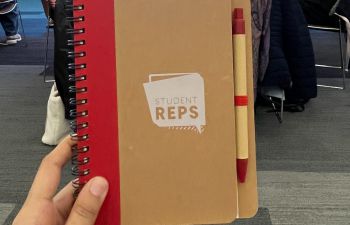 A hand holding up a notebook branded with the student rep scheme