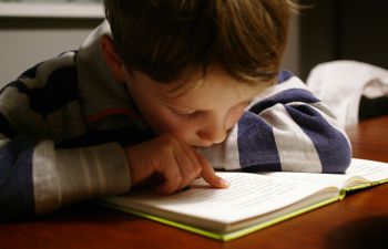 A child sits with their head in an open book, using their finger to trace words on the page