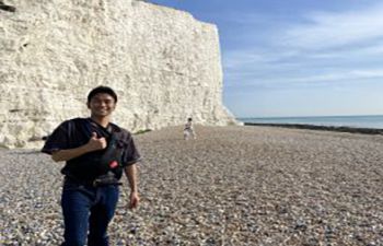 Student consultant Ryota is wearing a black t shirt and jeans, and giving the camera a thumbs-up. They are stood in front of a white cliff on a stony beach