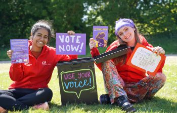 Two women wearing red Students' Union hoodies hold up signs and leaflets encouraging people to get involved.