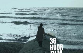 A still from 2023 entry to the competition. The scene is on the beach the sky is overcast and creates a grey feeling to the image. There is a person facing the sea wearing white. The text 