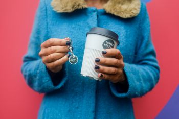 A woman holding the returnable cup in one hand and a token in the other, she's in a blue coat with a red background