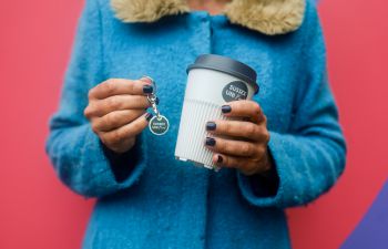 A woman holding the returnable cup in one hand and a token in the other, she's in a blue coat with a red background