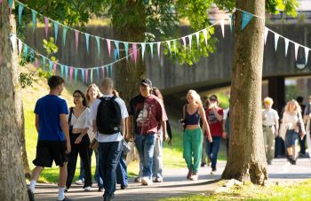 A group of University of Sussex students engaged in conversation, walking across campus on a sunny day under colourful bunting.