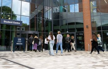 A diverse group of students is walking towards the Student Centre Building, with some entering through the revolving door and others walking in different directions outside.