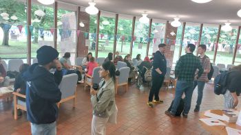 A photograph showing a room of people, who are chatting together in small groups. They look relaxed and happy. The room is brightly lit with large windows and comfortable seating.