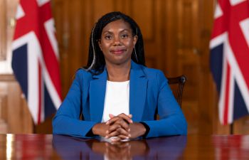 A photo of Kemi Badenoch at a desk with her hands on the surface, and two Union Jack flags in the background