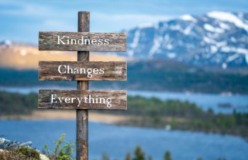 A wooden sign placed in the countryside, which says 'kindness changes everything'. There are mountains and a lake in the background