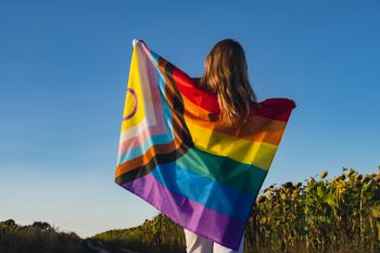 A woman faces away from camera with the progress pride flag across her shoulders