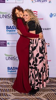 Hannah and Rebecca hug with joy in front of a screen promoting the sponsors of this high profile event