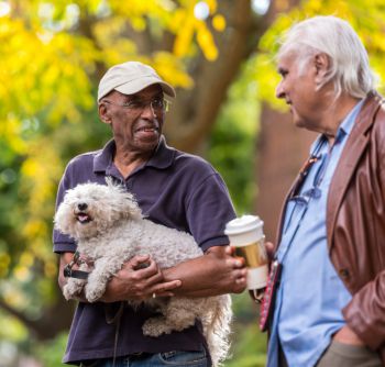 Two men in conversation outdoors, one holding a dog, with a tree in the background