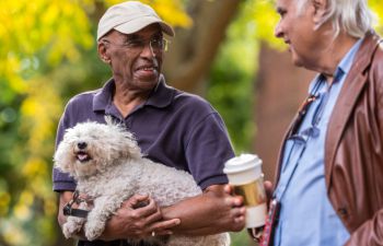 Two men in conversation outdoors, one holding a dog, with a tree in the background