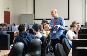 A lecturer wearing a red staff lanyard stands in the middle of a computer room with students talking